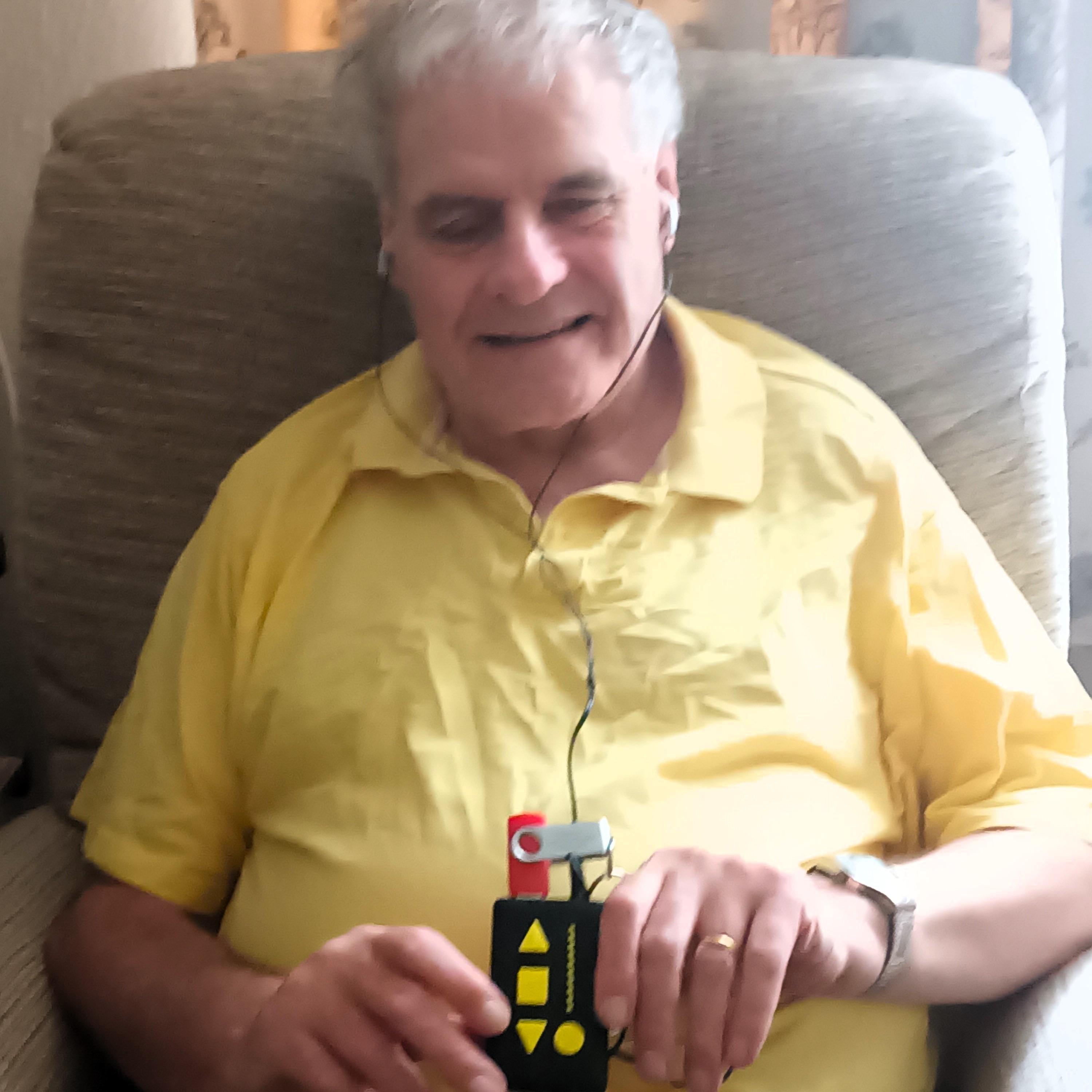 A grey haired man is sat in a beige arm chair, wearing a canary yellow shirt and holding a USB player. He has headphones in and is smiling.